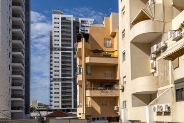 Mixed view of an Israeli residential district where aged stucco apartments with balconies and AC units stand beside modern glass high rises under a blue sky with scattered clouds