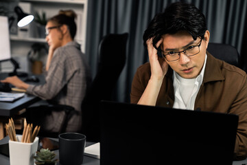 Businessman with stressful face working on laptop waiting email while coworker trading stock exchange investment at late night time at modern office, working on startup company project job. Infobahn.