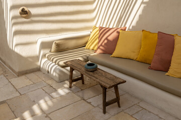 Inviting outdoor lounge with built in bench, soft cushions and colorful pillows under pergola slat shadows. Bright Mediterranean courtyard vibe ideal for hospitality lifestyle and design themes