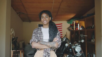 Confident woman standing in garage with motorcycle during daytime, showing passion for bikes and personal style