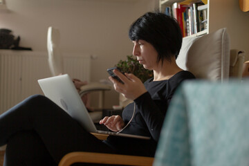 woman on armchair with laptop sipping. focused freelance editor reviews documents on laptop, holding warm mug, short hair, legs crossed, cozy livingroom with rug, shelf and plant, soft natural light,