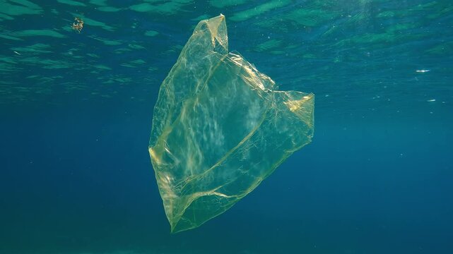 Old yellow plastic bag drifting below water reflected on surface in sunlight, Slow motion of disposable bag floating in blue water under sunbeams, Plastic pollution of Ocean 