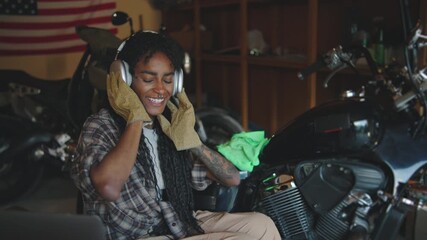 Young woman enjoys music while working on a motorcycle in a workshop with an American flag in the background - Powered by Adobe