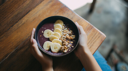 Hands hold a vibrant acai smoothie bowl topped with fresh banana slices and crunchy granola, a healthy and delicious breakfast served on a rustic wooden table.