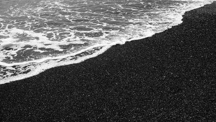 A black beach with a white wave. The beach is rocky and the water is choppy. The waves are crashing against the rocks.