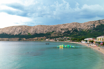 Obraz premium Scenic view of beach with inflatable water park and mountains under cloudy sky. Baška, Krk island, Croatia