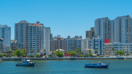 Bright, sunny view of a Brazilian city skyline, featuring modern apartment buildings overlooking a harbor with small fishing boats on the clear blue water