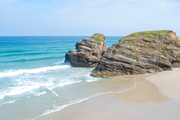 High-angle view of two massive sea stacks towering over pristine, sweeping sand at As Catedrais Beach, Spain. Tranquil blue water and rolling white waves complete the scenic coastal vista
