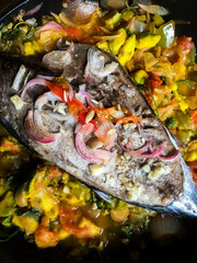 A close-up detail photo of grilled fish garnished with fresh onion, garlic, resting on a bed of mixed vegetables coated in curry sauce. Food suitable for culinary, restaurant, and healthy lifestyle
