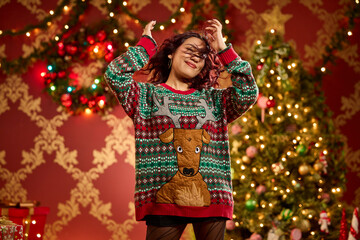 Woman wearing festive reindeer ugly Christmas sweater dancing in front of decorated holiday tree
