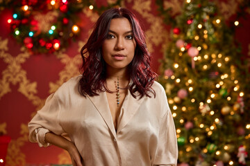 Woman with burgundy highlighted hair wearing cream blouse poses in front of illuminated Christmas tree