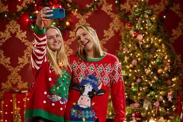 Two sisters taking a selfie while wearing festive Christmas sweaters in front of decorated tree