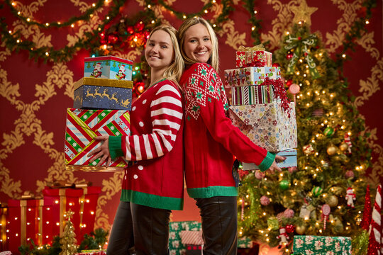 Two sisters in festive sweaters holding stacks of wrapped Christmas presents with holiday decorations