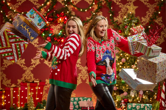 Two sisters in festive ugly Christmas sweaters celebrate with flying wrapped presents and holiday decorations