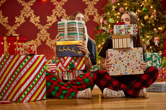 Two sisters in pajamas holding stacks of wrapped Christmas presents in front of decorated tree