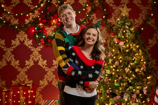 Couple wearing festive ugly Christmas sweaters pose together in front of decorated holiday tree