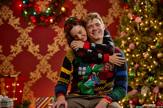 Happy couple embracing in matching ugly Christmas sweaters with festive holiday decorations and twinkling lights
