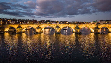 The view shows Charles Bridge in Prague during the evening. The bridge is lit up and reflects over...