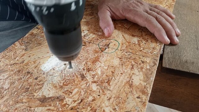 Close-up of a man drilling a hole in an OSB wood board with a cordless electric drill