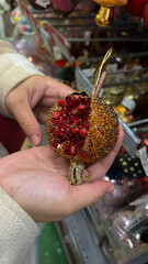 Vertical mobile photo. Female hand selects glass Christmas tree decoration, toy in shape of red pomegranate, bauble, for winter holiday in mass market shop, supermarket. New Year gift, eve, sale