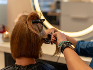 Fototapeta premium Hair styling with a straightener in a beauty salon, rear view, client sitting on a chair.