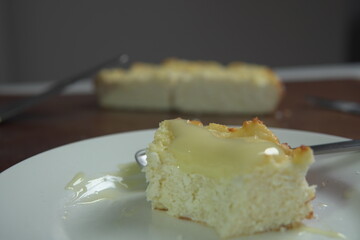 appetizing slice of homemade cottage cheese casserole topped with flowing condensed milk on white plate with fork bite and whole casserole in soft background