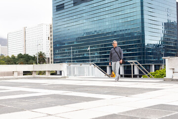 Senior man walking across tiled terrace by railing holding black bag, yellow umbrella, copy space