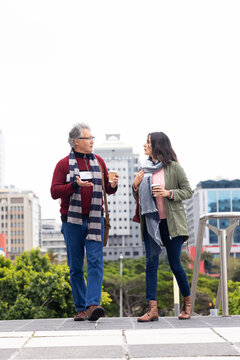 Diverse colleagues walking on urban terrace wearing green parka and red sweater holding coffee cups