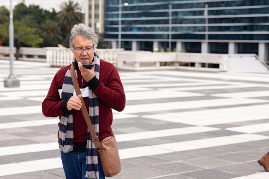 Senior man standing in urban plaza speaking into smartphone with striped scarf and leather bag