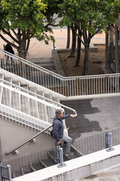 Senior male standing on metal stair landing in outdoor plaza pointing forward with backpack