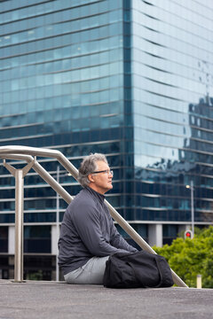 Senior man sitting on platform beside railing at office plaza with duffel bag, copy space