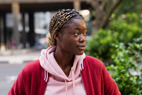 African American woman standing on city sidewalk wearing pink hoodie and red jacket gazing right