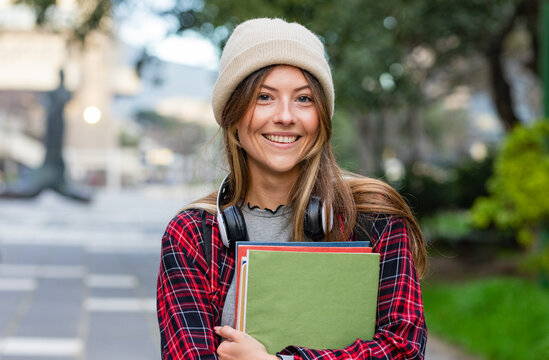 Female student standing on campus walkway wearing plaid shirt holding notebooks and headphones