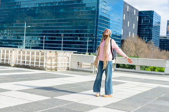 African American woman opening arms tilting head on plaza by railing with headphones and backpack