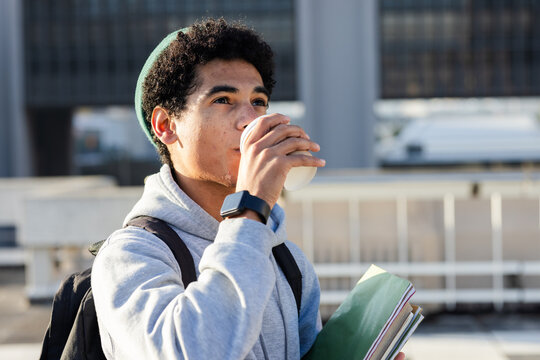 Male student standing on transit platform wearing backpack, holding notebooks and sipping coffee