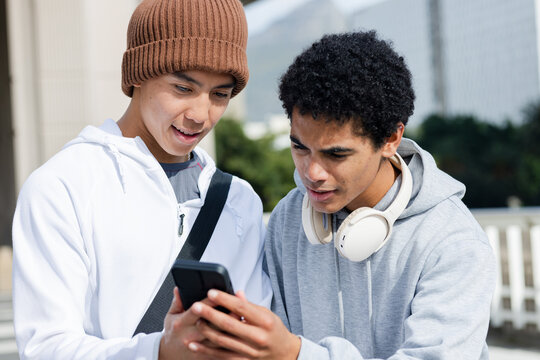 Diverse male friends checking smartphone screen on urban terrace with crossbody bag and headphones