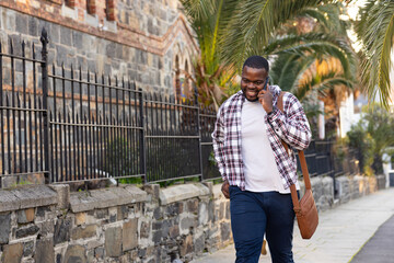 African American man walking past stone building using smartphone with messenger bag, copy space