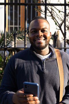 African American male standing by iron fence holding smartphone with shoulder bag strap