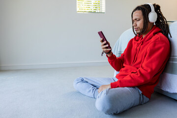 African American man sitting at home in red hoodie using phone with headphones, copy space