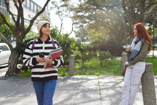 Diverse female friends walking campus path carrying folders and checking smartphones, copy space
