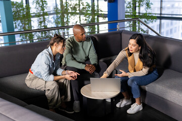 Diverse coworkers collaborating around silver laptop on sectional sofa at office lounge