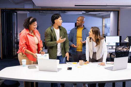 Diverse coworkers standing around table in office discussing chart data laptops and smartphone