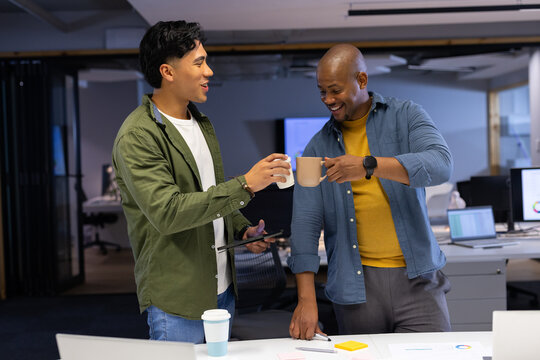 Diverse male coworkers raising ceramic mugs and reviewing tablet at office desk with charts
