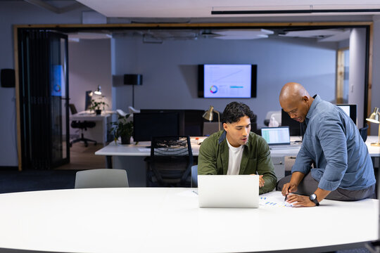 Diverse male coworkers collaborating on data in modern office with silver laptop and printed charts