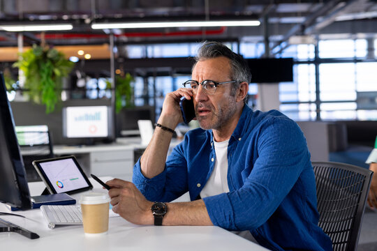Mature man sitting at desk in open plan office holding smartphone and pointing at tablet chart
