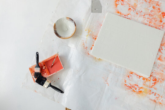 Blank primed canvas resting on splattered drop cloth, with coral paint tray, brushes, bowl, scraper