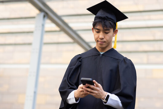 Asian man wearing mortarboard and gown checking smartphone in campus courtyard with smartwatch