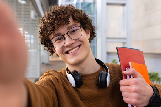 Male student holding notebooks and smartphone taking selfie in university corridor with headphones