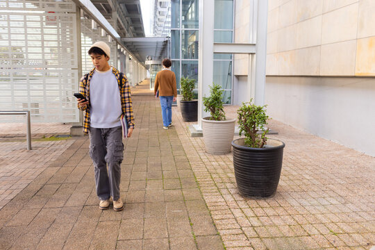 Diverse men walking along paved walkway beside glass building carrying smartphone, backpack