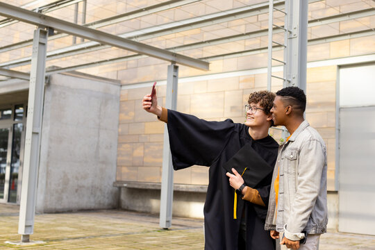 Diverse male friends celebrating graduation on campus with smartphone and mortarboard, copy space